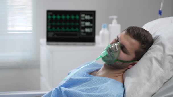 Caucasian man with oxygen mask on lying in bed with white linen, sleeping disturbingly, moving head. Portrait of a man in an oxygen mask who lies on a bed in a hospital. — Video