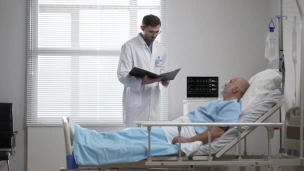 Young doctor in glasses talking to senior man resting in hospital bed. Male RN assisting elderly male patient lying down in bed. Friendly male Doctor Checks on the Sick Male Patient Lying in Bed — Vídeos de Stock