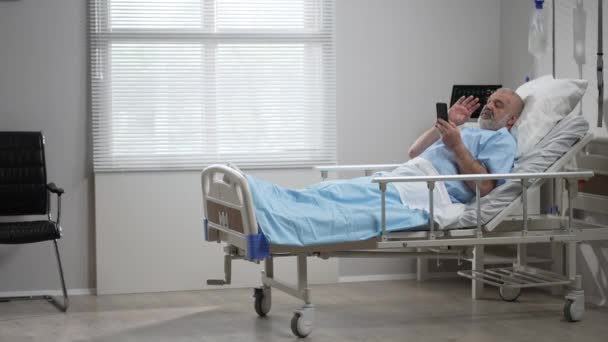 Hospital Ward: senior man Resting in Bed uses Smartphone for Video Call Conference Talk with Family and Friends. video call in hospital room — Video Stock