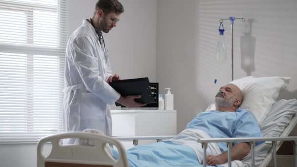 Young doctor talking to senior man resting in hospital bed. Male RN assisting elderly male patient lying down in bed. Friendly male Doctor Checks on the Sick Male Patient Lying in Bed — Stock Video