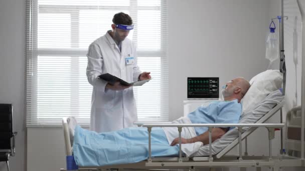 Young doctor in protective mask screen talking to senior man resting in hospital bed. Male assisting elderly male patient lying down in bed. Male Doctor Checks on the Sick Male Patient Lying in Bed — Vídeo de Stock