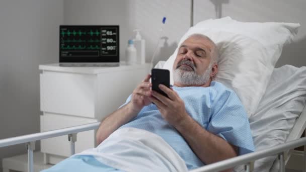 Hospital Ward: senior man Resting in Bed uses Smartphone for Video Call Conference Talk with Family and Friends. video call in hospital room — Vídeo de Stock