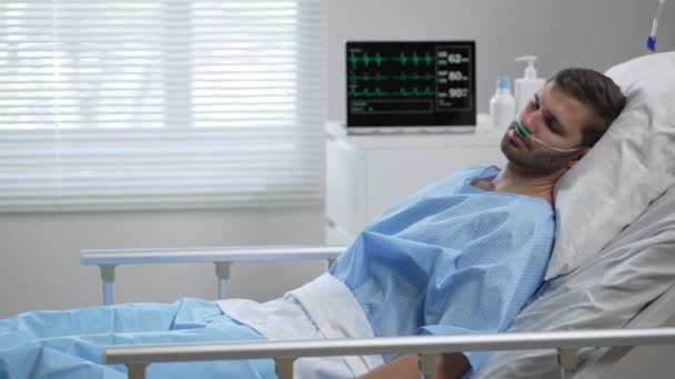 A young man lies on a bloodied hospital and opens his eyes. apparatus for monitoring heart, oxygen and pulse. A young patient of the clinic in a coma. Regain consciousness — Vídeo de Stock