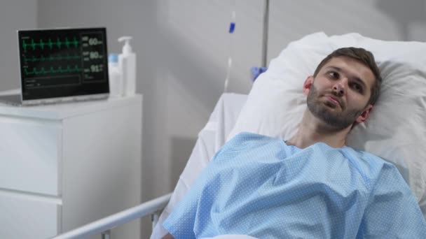 A young man lies on a bloodied hospital and opens his eyes. apparatus for monitoring heart, oxygen and pulse. A young patient of the clinic in a coma. Regain consciousness — Stock videók