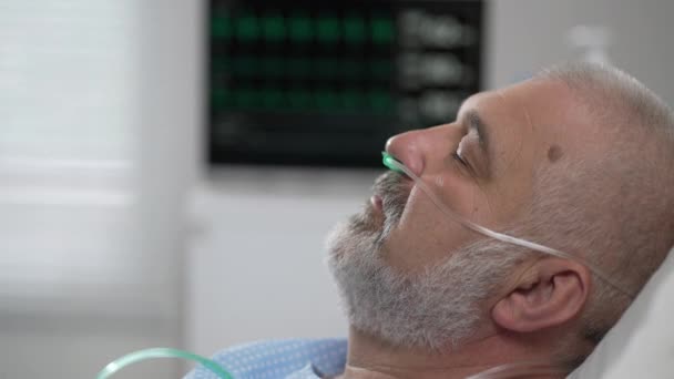 The face of an elderly man lying on a bed in a hospital, unconscious, asleep. The ECG machine shows vital signs — Stockvideo