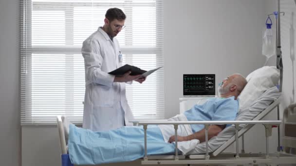 Young doctor talking to senior man resting in hospital bed. Male RN assisting elderly male patient lying down in bed. Friendly male Doctor Checks on the Sick Male Patient Lying in Bed — Video