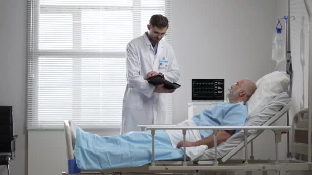 Young doctor in glasses talking to senior man resting in hospital bed. Male RN assisting elderly male patient lying down in bed. Friendly male Doctor Checks on the Sick Male Patient Lying in Bed — ストック動画