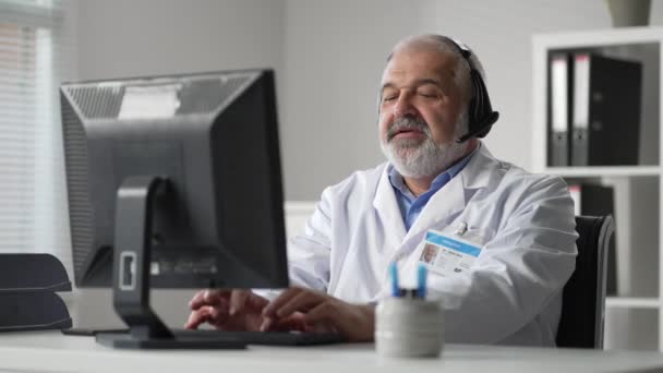 Médico masculino gris de forma remota las comunicaciones de vídeo con el cliente y los registros de la historia clínica en el ordenador — Vídeo de stock