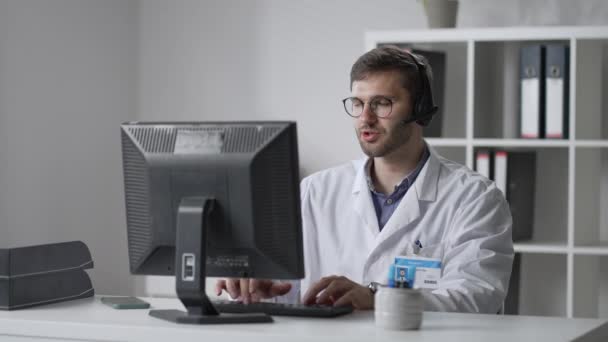 Beyaz önlüklü erkek pratisyen doktor muayenehanesinde masasında oturuyor ve hastaların tıbbi geçmişini okurken bilgisayar faresi kaydırıyor. — Stok video