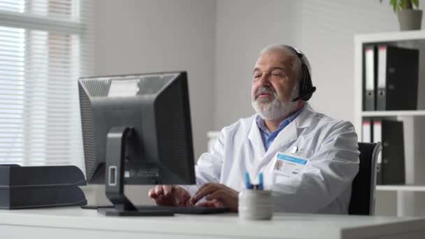 Cinza médico masculino remotamente comunicações de vídeo com o cliente e registra histórico médico no computador — Vídeo de Stock