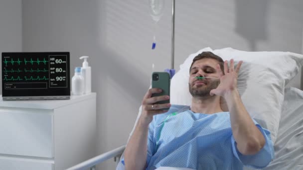 Молодой пациент клиники лежит на больничной койке и звонит по видеосвязи с помощью мобильного телефона своей семье и друзьям. — стоковое видео