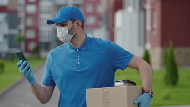 El repartidor masculino en una gorra y una máscara protectora y guantes va con una caja en las manos y lleva un paquete al cliente. Entrega de pedidos en línea a domicilio. — Vídeos de Stock