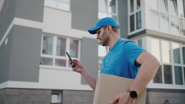 A male mail carrier with round glasses carries boxes for delivery to customers and looks at his mobile phone. Search through the mobile phone for the delivery address. Payment terminal. — Stock Video