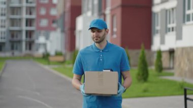 Mavi teslimatçı kargodan önce müşteriye bir paket gönderir. 4k çözünürlük ve yavaş çekim. Şehir caddesinde elinde paket olan bir erkek postacı. Posta konsepti. Teslimat servisi.