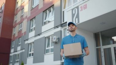 Mavi teslimatçı kargodan önce müşteriye bir paket gönderir. 4k çözünürlük ve yavaş çekim. Şehir caddesinde elinde paket olan bir erkek postacı. Posta konsepti. Teslimat servisi.