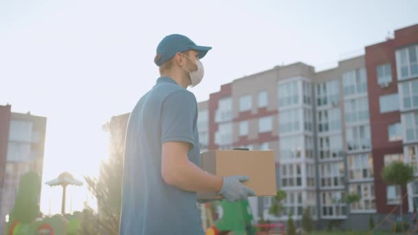 Şapkalı, koruyucu maskeli ve eldivenli erkek teslimatçı elinde bir kutuyla müşteriye bir paket taşır. Çevrimiçi ev siparişleri teslim ediliyor. — Stok video