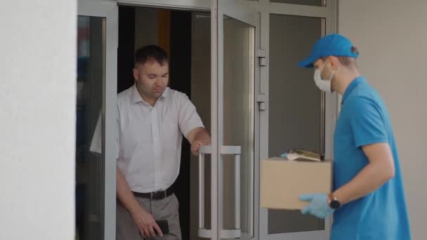 The male deliveryman in masks and protective gloves carries a parcel to the customer with a terminal for contactless payment by mobile phone or NFC card. — Stock Video