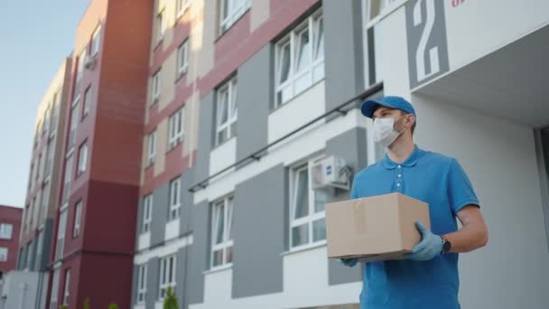 Seorang kurir tukang pos muda dengan topeng pelindung dan sarung tangan mengirimkan paket langsung ke rumah pelanggan dengan aman. Konsep kurir, pengiriman rumah, pengiriman e-commerce, virus, covid — Stok Video