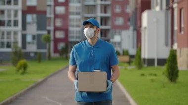 Şapkalı, koruyucu maskeli ve eldivenli erkek teslimatçı elinde bir kutuyla müşteriye bir paket taşır. Çevrimiçi ev siparişleri teslim ediliyor.