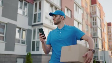 Yuvarlak gözlüklü bir erkek postacı müşterilere teslim etmek için kutular taşır ve cep telefonuna bakar. Cep telefonundan teslimat adresini ara. Ödeme terminali.