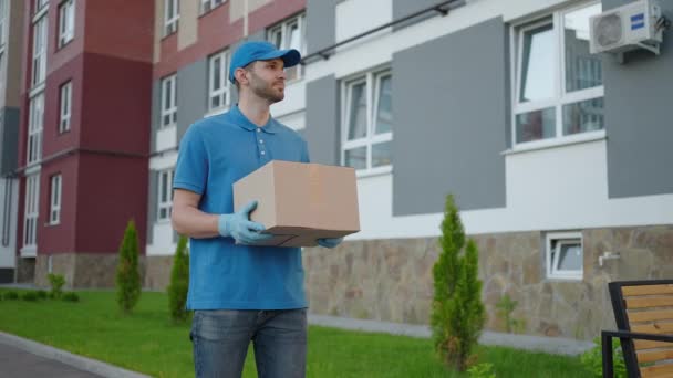 Um homem de entrega em uma camiseta azul e boné carrega uma caixa de papelão na cidade olhando em volta em busca do endereço do entregador. Entregador vai com uma caixa em câmera lenta — Vídeo de Stock