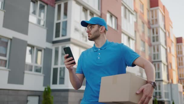 Yuvarlak gözlüklü bir erkek postacı müşterilere teslim etmek için kutular taşır ve cep telefonuna bakar. Cep telefonundan teslimat adresini ara. Ödeme terminali. — Stok video