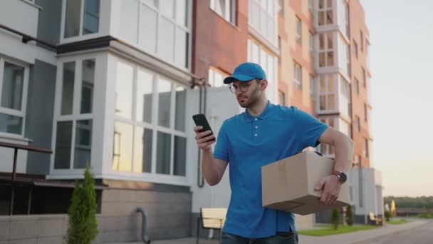 Un facteur masculin avec des lunettes rondes transporte des boîtes pour la livraison aux clients et regarde son téléphone mobile. Rechercher dans le téléphone portable l'adresse de livraison. Terminal de paiement. — Video