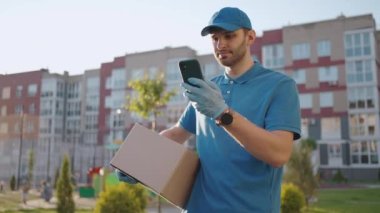 The postman with glasses carries the parcel and looks at the delivery address via mobile phone. search for the address of the delivery customer. Delivery guy with a cap and a box