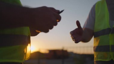 Construction manager and workers shaking hands on construction site. Builder man with a tablet and a man inspector in white helmets shake hands at sunset standing. Symbol of agreement successful work.