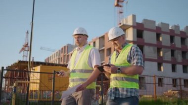 Two engineers discussing project on a construction site, a worker wearing a helmet during the sunset
