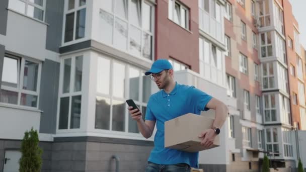 白人年轻貌美的男子，头戴蓝色帽子的送货工人一边走在街上，一边提着纸盒，一边用智能手机，寻找路线。有包裹窃听和电话发短信的男性信使. — 图库视频影像