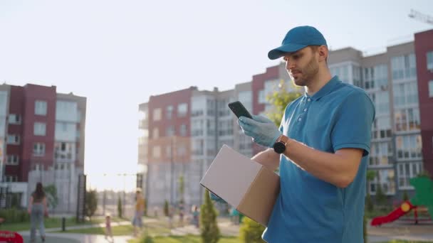 Listonosz w okularach niesie paczkę i patrzy na adres dostawy przez telefon komórkowy. poszukiwanie adresu odbiorcy dostawy. Dostawca z czapką i pudełkiem — Wideo stockowe