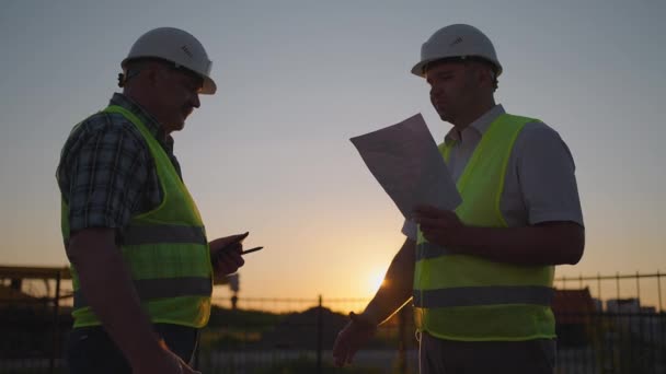Construction manager and workers shaking hands on construction site. Builder man with a tablet and a man inspector in white helmets shake hands at sunset standing. Symbol of agreement successful work. — Stok video