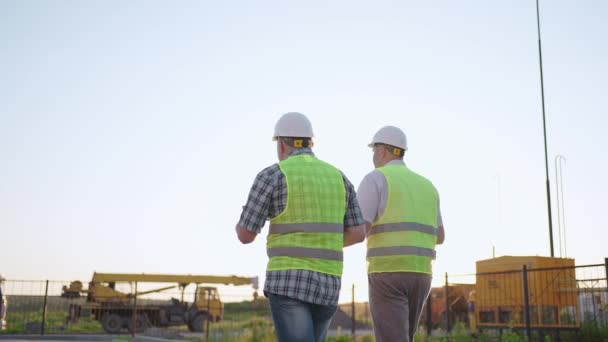 Pas-up dvou mužů středního věku stavitelé nosí bezpečnostní oblečení, stojí na staveništi, muž pomocí vysílačky, jeho kolega drží papír s plánem projektu — Stock video