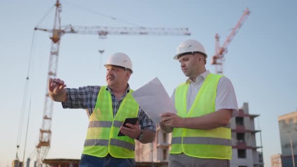 Портрет двух строителей, стоящих на строительной площадке. Два строителя с чертежами стоят на фоне строящихся зданий в шлемах и жилетах — стоковое видео