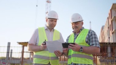 Portrait of two builders standing at building site. Two builders with drawings standing on the background of buildings under construction in helmets and vests