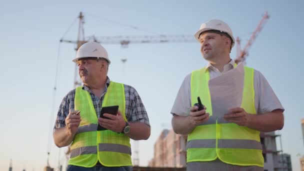 Cintura de dois construtores masculinos de meia-idade vestindo roupas de segurança, de pé no local de construção, homem usando walkie-talkie, seu colega segurando papel com plano de projeto — Vídeo de Stock