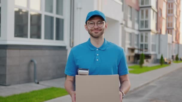 Un repartidor feliz con una caja en las manos mira a la cámara y sonríe. Retrato de un repartidor con una caja en cámara lenta — Vídeo de stock