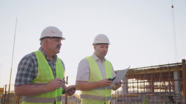 Tour de taille de deux constructeurs masculins d'âge moyen portant des vêtements de sécurité, debout sur le chantier, homme utilisant talkie-walkie, son collègue tenant du papier avec le plan de projet — Video