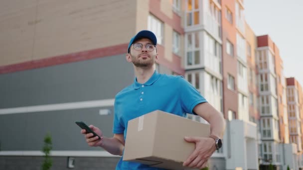 O carteiro com óculos carrega o pacote e olha para o endereço de entrega via celular. procurar o endereço do cliente de entrega. Entrega cara com um boné e uma caixa — Vídeo de Stock