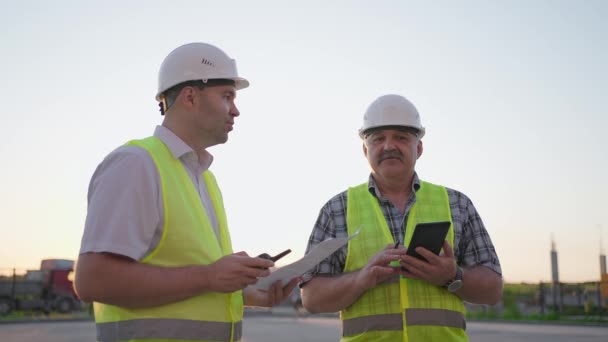 Tour de taille de deux constructeurs masculins d'âge moyen portant des vêtements de sécurité, debout sur le chantier, homme utilisant talkie-walkie, son collègue tenant du papier avec le plan de projet — Video