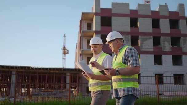 Tour de taille de deux constructeurs masculins d'âge moyen portant des vêtements de sécurité, debout sur le chantier, homme utilisant talkie-walkie, son collègue tenant du papier avec le plan de projet — Video