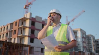 Foreman in helmet and vest talking on walkie-talkie with builders, standing at construction site, tracking shot. building expert engineers speaking using a radio with some builders , amazing sunlight.