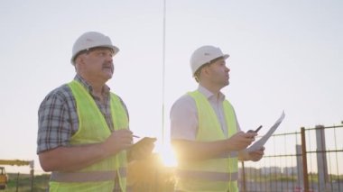 Portrait of two builders standing at building site. Two builders with drawings standing on the background of buildings under construction in helmets and vests