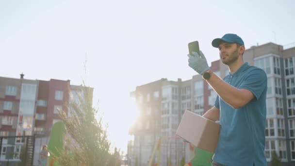 El cartero con gafas lleva el paquete y mira la dirección de entrega a través del teléfono móvil. buscar la dirección del cliente de entrega. Repartidor con gorra y caja — Vídeos de Stock