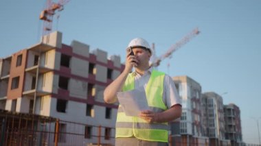 Foreman in helmet and vest talking on walkie-talkie with builders, standing at construction site, tracking shot. building expert engineers speaking using a radio with some builders , amazing sunlight.