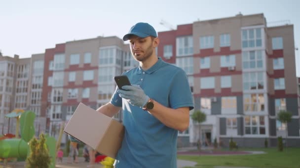 眼鏡をかけた郵便配達員が小包を運び、携帯電話で配達先を見ている。お客様の住所を検索してください。キャップと箱を持った配達人 — ストック動画