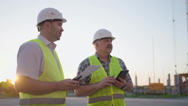 Dva inženýři diskutují o projektu na staveništi, dělník v helmě během západu slunce — Stock video