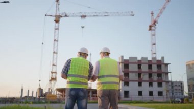 Orta yaşlı, güvenli kıyafetler giyen, inşaat alanında duran, telsiz kullanan, iş arkadaşı elinde proje planı olan iki erkek müteahhit.