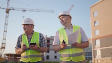 Portrait of two builders standing at building site. Two builders with drawings standing on the background of buildings under construction in helmets and vests